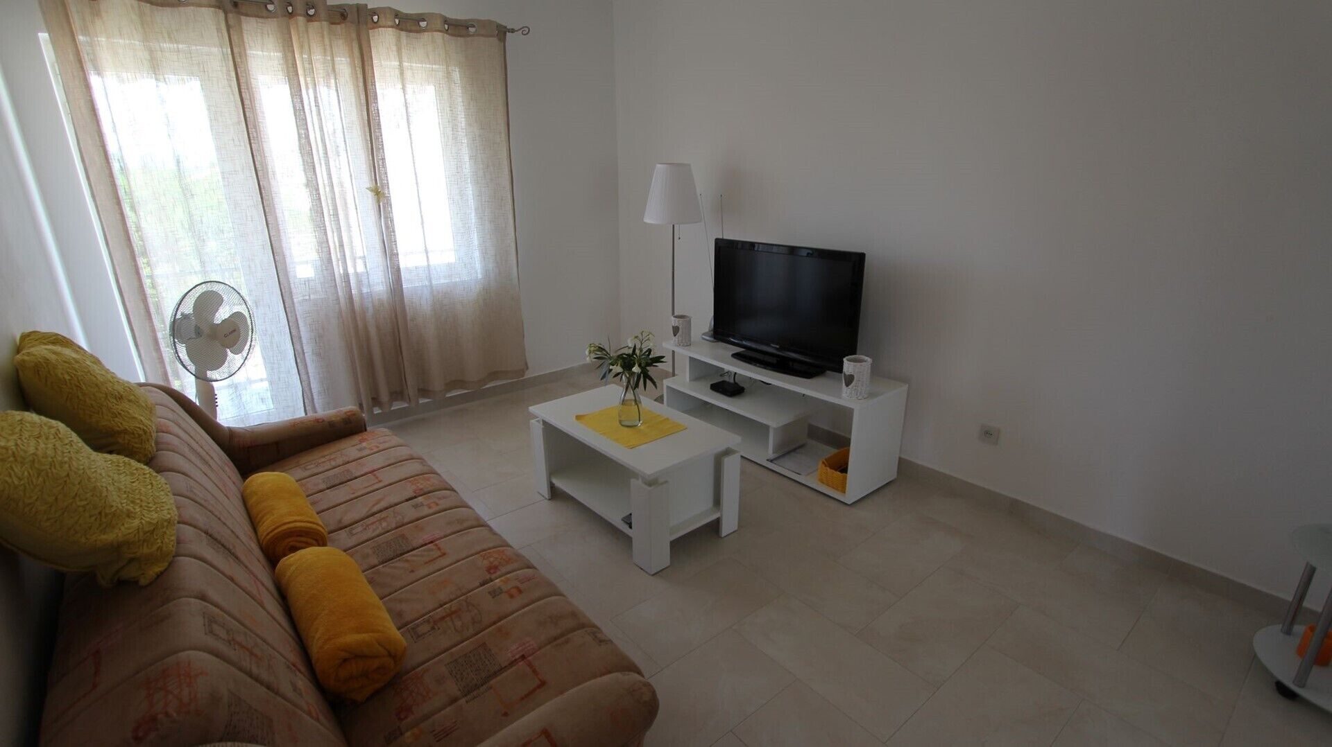 A living room with a sofa, a TV on a stand, a small table with a plant, and a fan near the window.