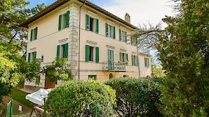 A two-story building with green shutters, a balcony with a wrought iron railing, and a garden area with a lawn and bushes. - Elegant villa on Lake Trasimeno - Fiordaliso (Ranciano)