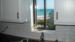 Una cocina con armarios blancos, una ventana con vistas al océano y una casa de playa, y un fregadero.
