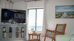 Una sala de estar con televisor, un sillón de balancín de madera, una mesita y vista al océano a través de un ventanal grande.