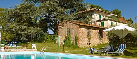A swimming pool with lounge chairs and umbrellas, a stone building with a garden, and a large tree.