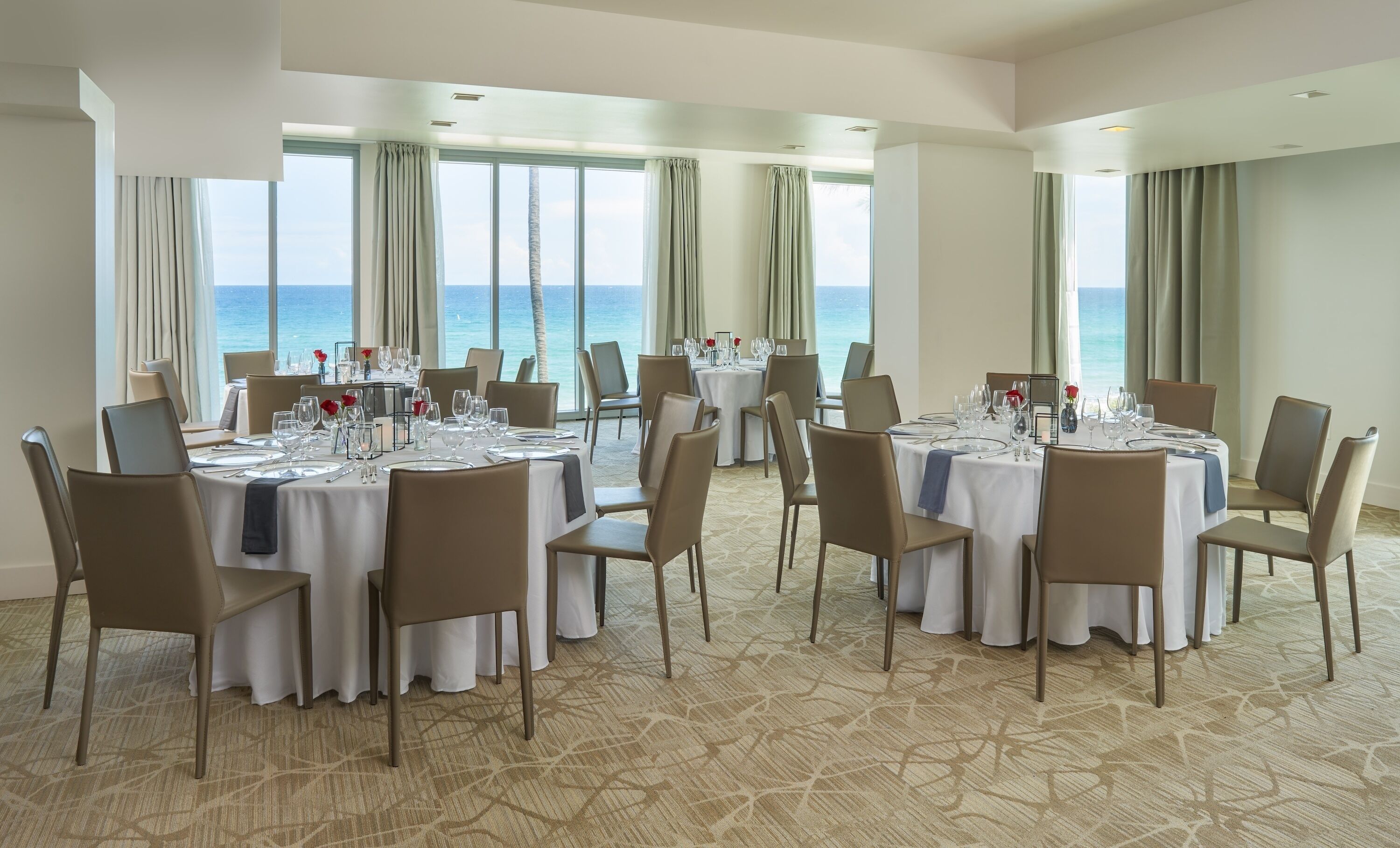 Un espace repas avec des tables rondes disposées pour accueillir six personnes, de grandes fenêtres avec des rideaux et une vue sur l’océan.