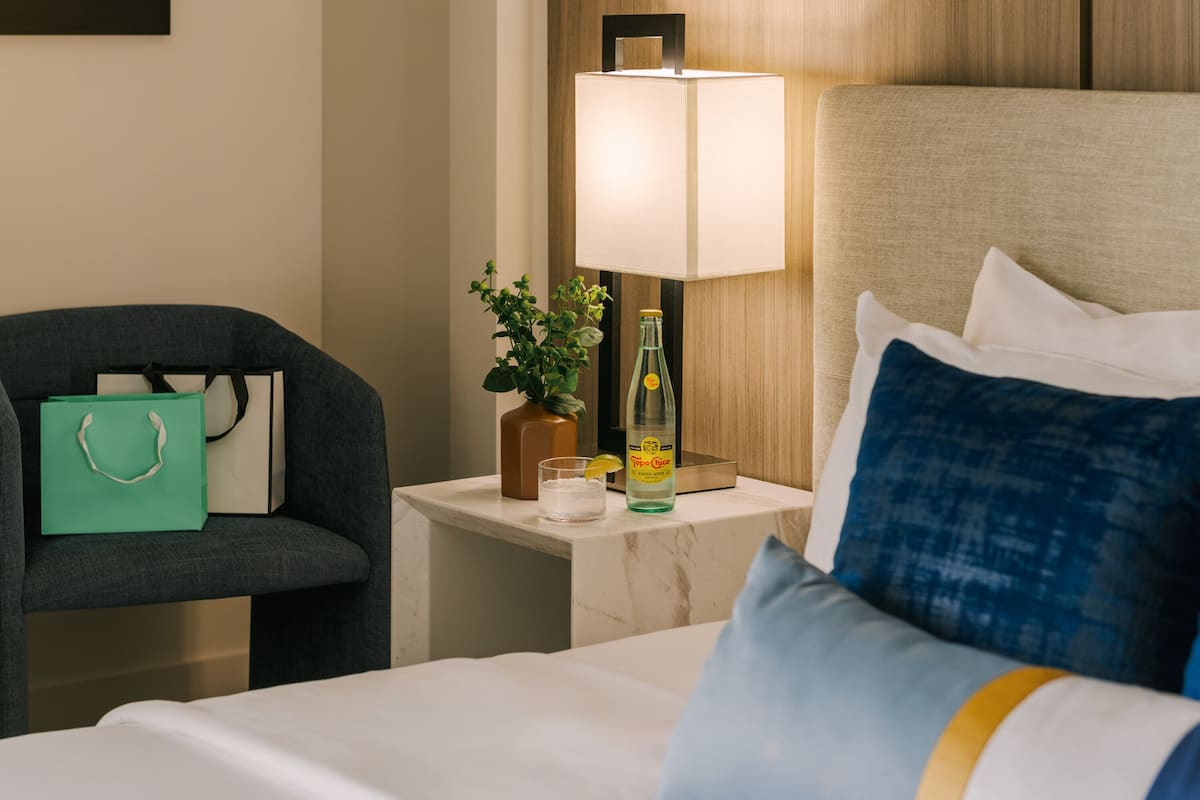 A hotel room with a bed, a nightstand, a lamp, a green bag, a glass of water, and a bottle of hand sanitizer.