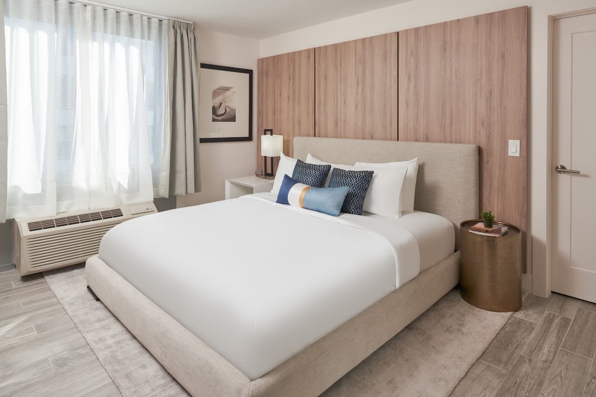 A modern hotel room with a large bed, a bedside table with a lamp, a small plant, and a wooden panel wall.