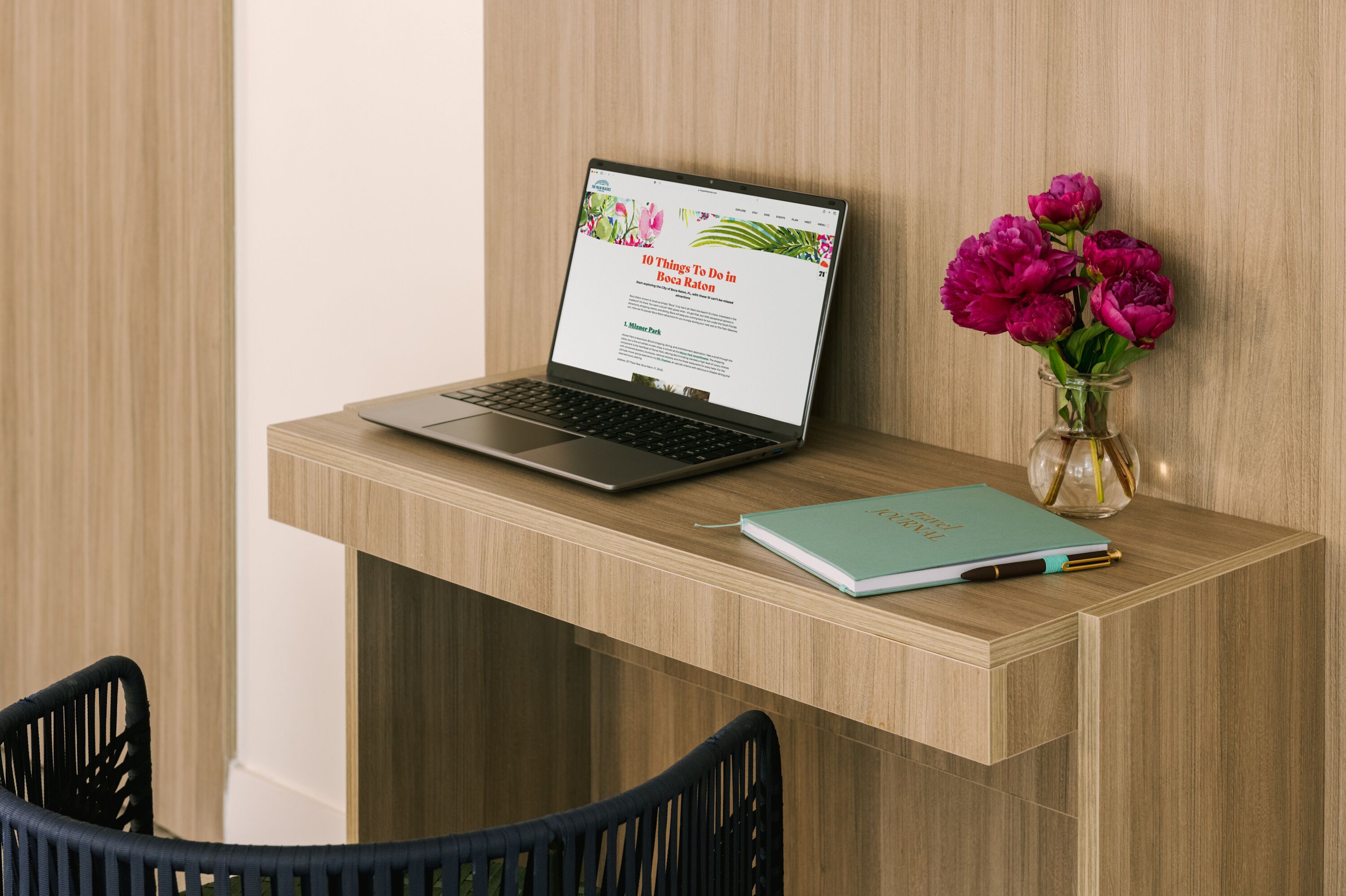 A laptop on a wooden desk with a notebook and a vase of flowers.