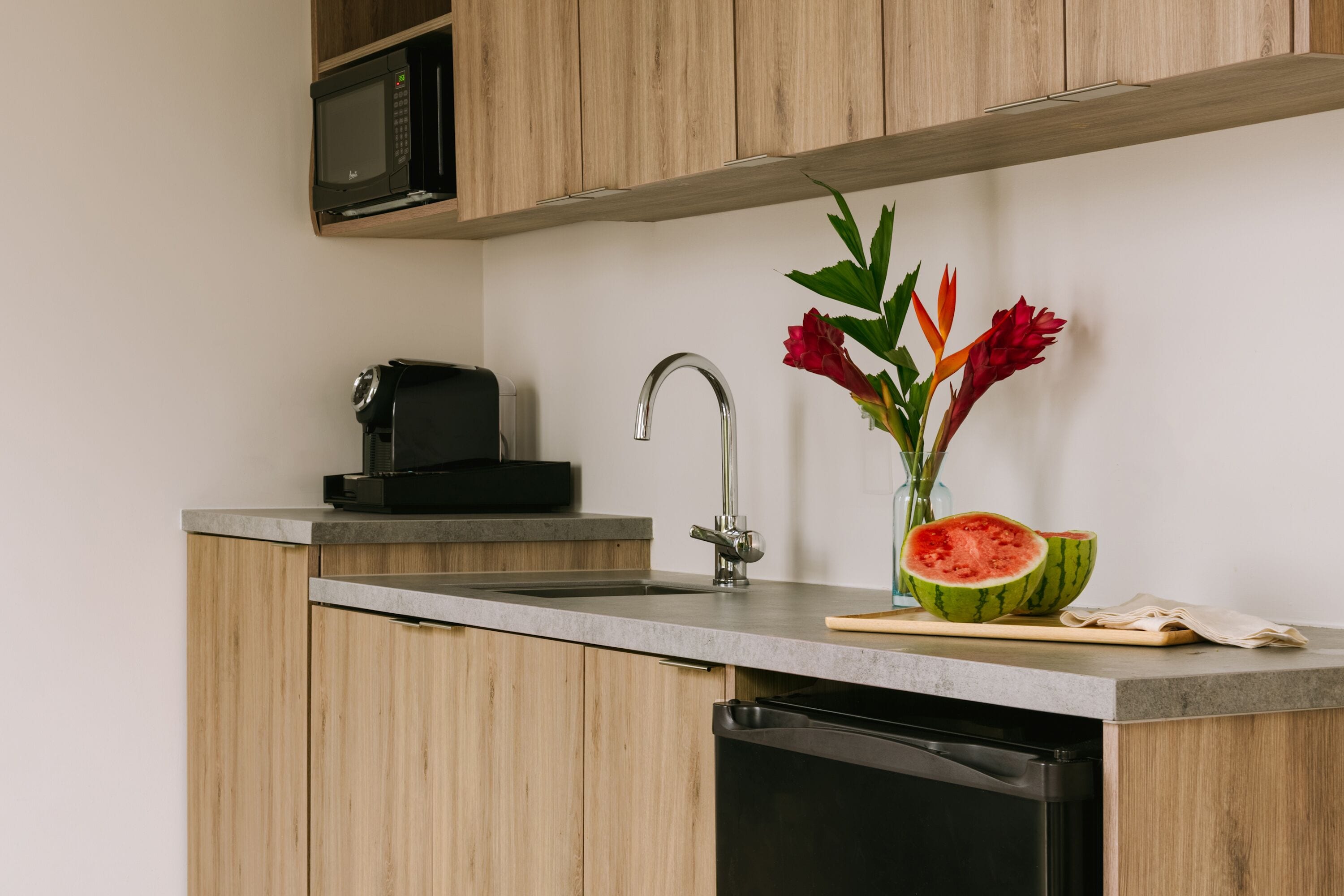 A modern kitchen with wooden cabinets, a built-in microwave, a stainless steel sink, and a vase with flowers and a watermelon slice.