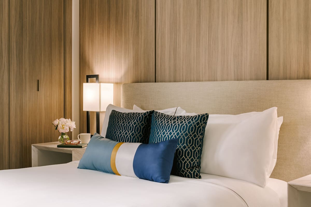 A hotel room with a bed, pillows, a nightstand, a lamp, and a vase with flowers.