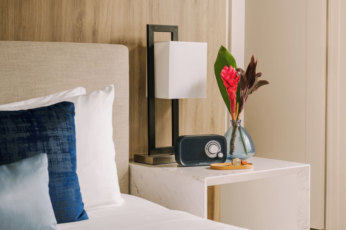 A neatly made bed with a white and blue pillow, a bedside table with a radio, a vase with flowers, and a wall-mounted lamp.