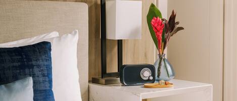 A neatly made bed with a white and blue pillow, a bedside table with a radio, a vase with flowers, and a wall-mounted lamp.