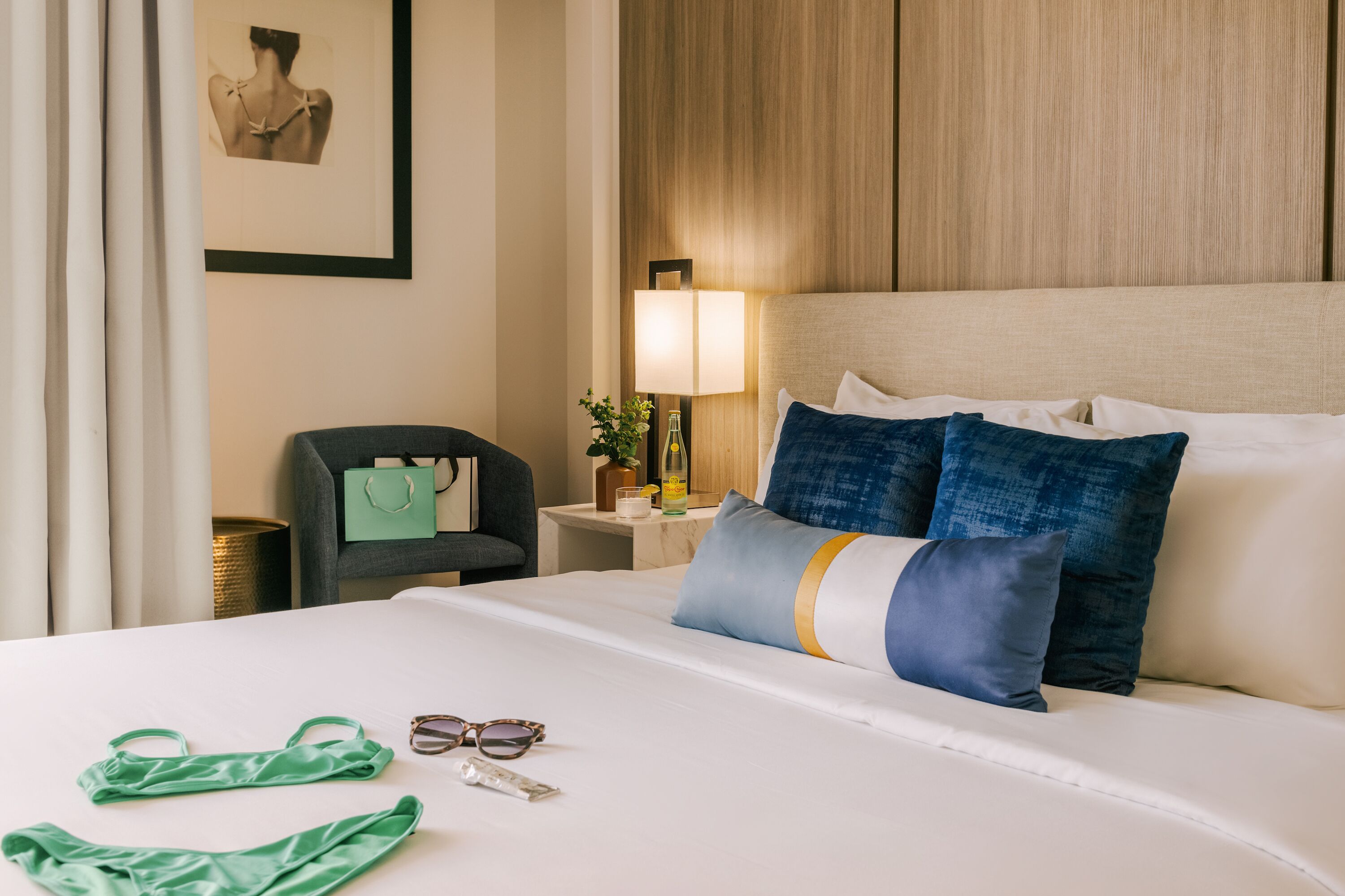 A hotel room with a neatly made bed, a bedside table with a lamp, a chair, and a framed picture on the wall.