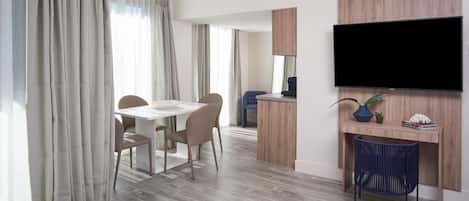 A modern hotel room with a dining area, a TV mounted on the wall, and a small desk with a plant.