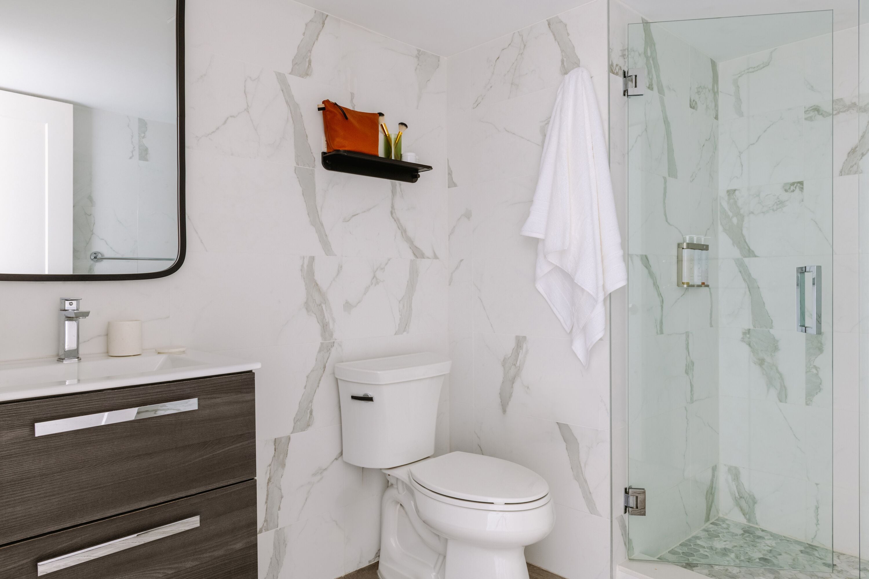 A bathroom with a marble wall, a white toilet, a dark wood vanity, a glass shower enclosure, and a towel hanging on the wall.