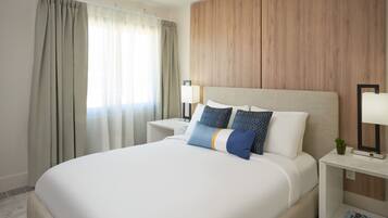Habitación de hotel con una cama grande, cabecera de madera, dos mesitas de noche con lámparas y una ventana con cortinas.