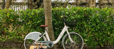 En hvid cykel med kurv og taske, parkeret på græsset ved en hæk.