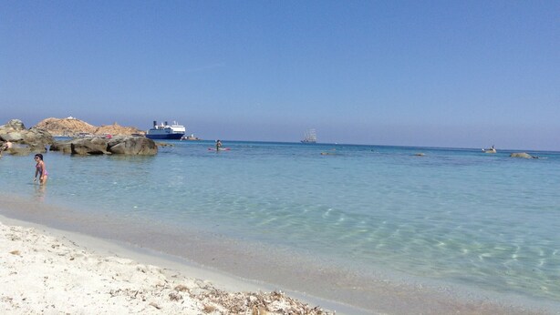 Una playa con agua cristalina, rocas y un barco a lo lejos.