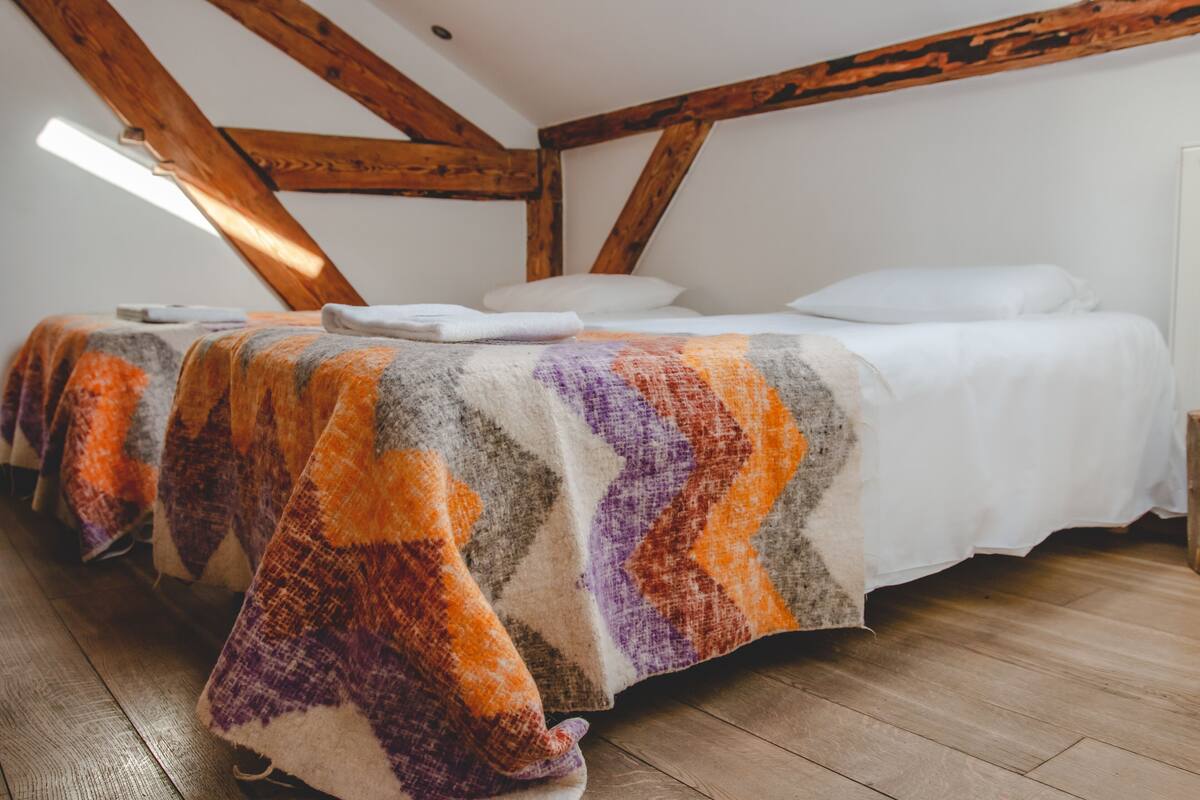 A cozy attic bedroom with exposed wooden beams, a wooden floor, and a colorful blanket on the bed.