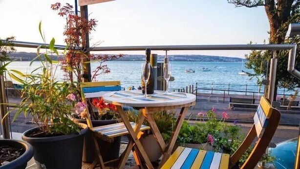 Een balkon met een ronde tafel en stoelen, potplanten en uitzicht op het water.