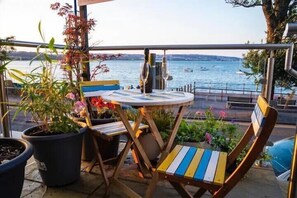 Een balkon met een ronde tafel en stoelen, potplanten en uitzicht op het water.
