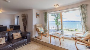 Een woonkamer met een leren bank, een houten salontafel en twee schommelstoelen. Een groot raam biedt uitzicht op zee.