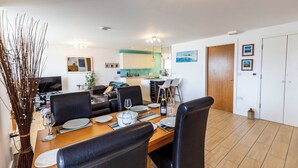 Een eetruimte met een houten tafel voor vier personen, omringd door zwarte stoelen. Achtergrond is een keuken met witte kasten en een blauwe accentmuur.