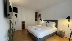 A neatly arranged bedroom with a large bed, built-in wardrobe, bedside lamps, and a small bedside table.