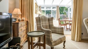 A living room with a television, a wooden cabinet, a plaid armchair, a small round table, and a dining area with chairs and a table. - High Trees Blenheim View | Woodstock Holiday Let | 3 Beds & Sleeps 5 (Woodstock)