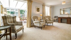 A spacious living room with a large window, two armchairs, a sofa, a wooden desk, and a lamp. - High Trees Blenheim View | Woodstock Holiday Let | 3 Beds & Sleeps 5 (Woodstock)