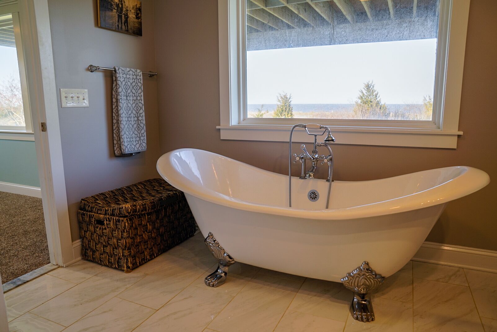 Un baño con una bañera independiente, un pequeño cofre de mimbre, un perchero y una ventana con vistas a árboles y una masa de agua.