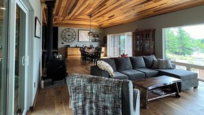 Een ruime woonkamer met een houten plafond, een grijze bank, een salontafel en een eethoek met een grote klok en een open haard.