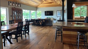 Een ruime woonkamer met een houten plafond, een eettafel met stoelen, een bank, een keuken met een groot raam, en een aan de muur bevestigde televisie.