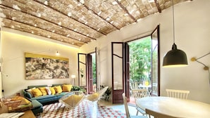 A spacious living area with a checkered floor, a sofa with yellow cushions, a dining table with chairs, and a large window offering a view of greenery outside. - Tyche Apartment Barcelona (Barcelona)