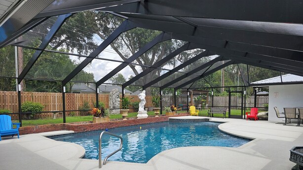 A covered outdoor swimming pool with a metal ladder, surrounded by a wooden fence, and featuring outdoor furniture and decorative plants. - Great quiet and comfortable, South Central Lakeland location. (Lakeland)