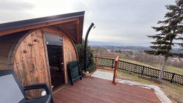 A wooden cabin with an open door, a black chair, a small table, and a view of a town and hills.