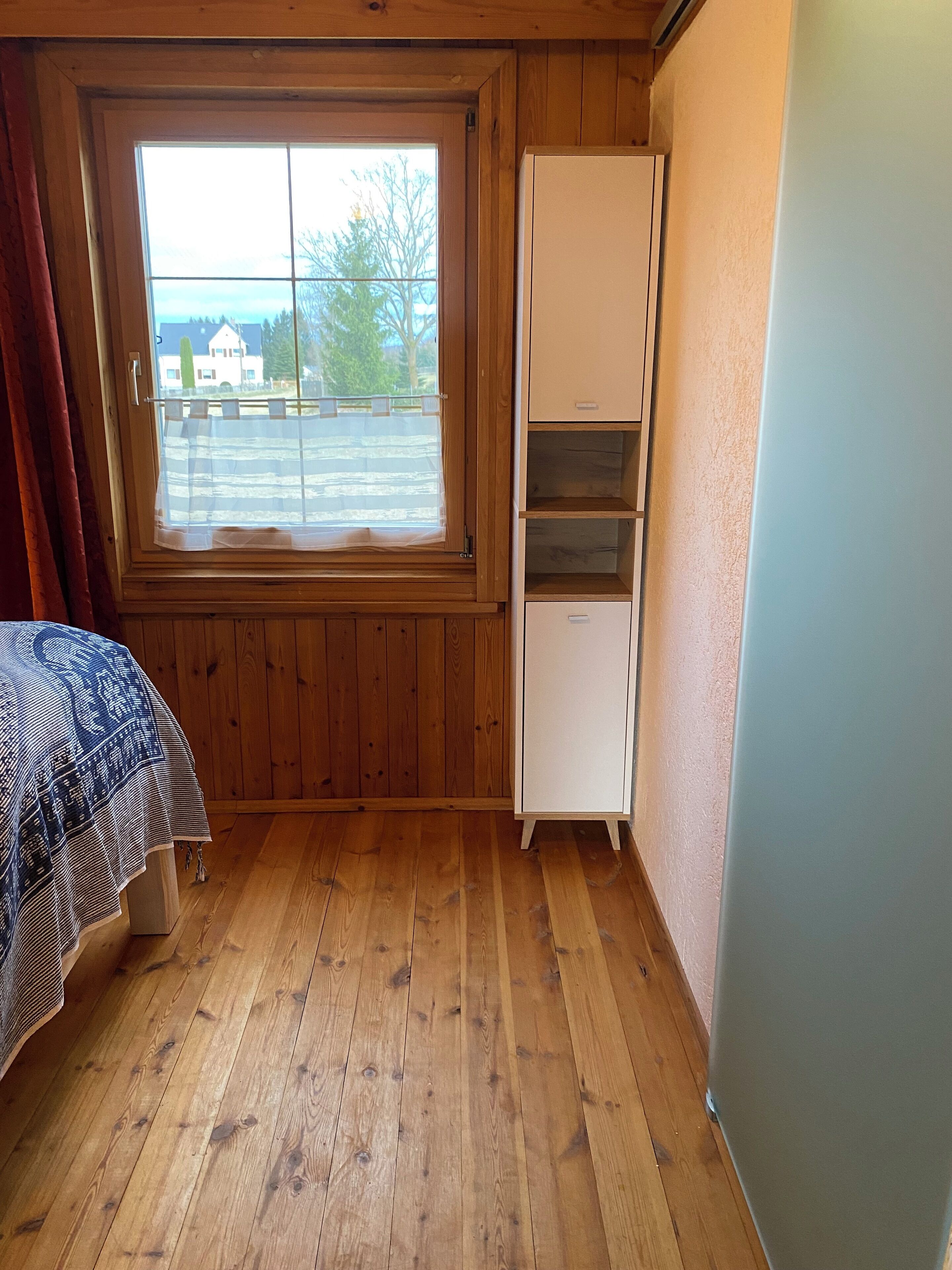 Ett rum med träväggar, ett fönster med utsikt över ett hus och träd, en säng med ett mönstrat överkast och ett vitt skåp.