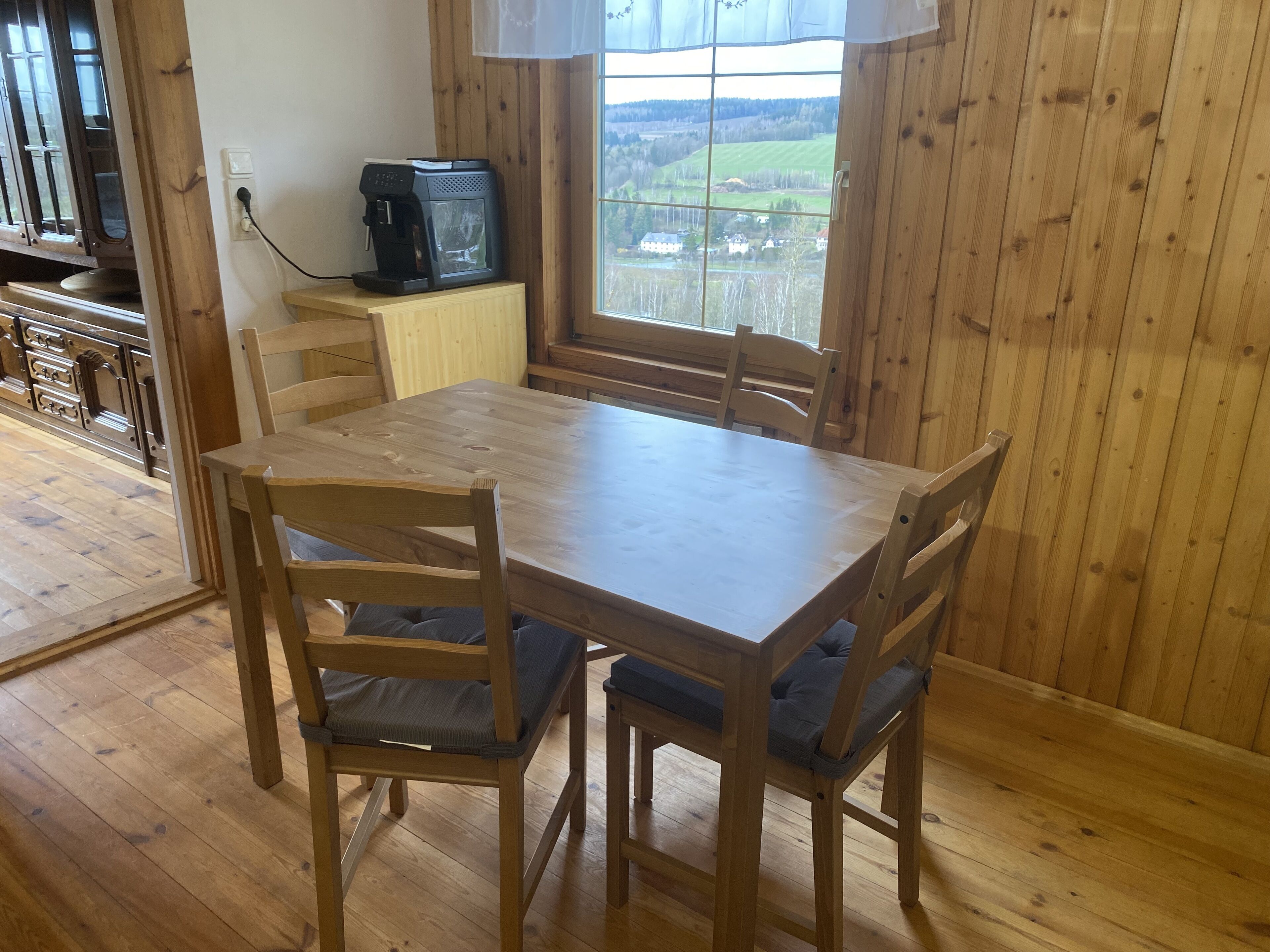 A wooden dining table with four chairs, a coffee maker on a small wooden cabinet, and a window with a view of a rural landscape.