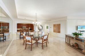 Un vaste espace dédié à la salle à manger, avec une table en verre et des chaises en bois, une cuisine dotée de meubles en bois, un lustre et un téléviseur posé sur une console.