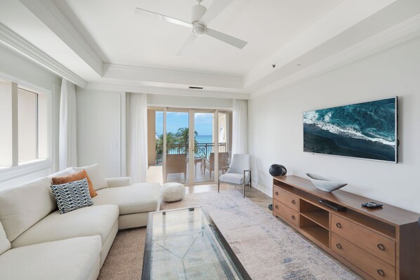 A spacious living room with a large window, a ceiling fan, a flat-screen TV, and a view of the ocean.