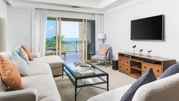A spacious living room with a large sofa, a coffee table, a flat-screen TV, and a balcony with a view of the ocean. - Sophisticated Ocean View Residence located at The Ritz-Carlton (Seven Mile Beach)