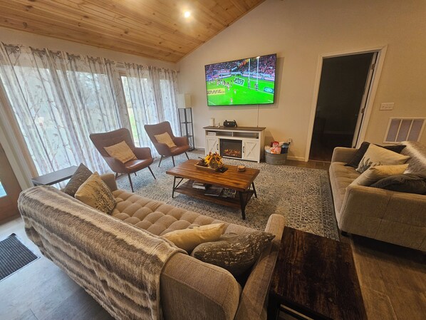 A cozy living room with a fireplace, comfortable seating, and a large TV displaying a sports game. - Relax in our large 4b/2b ranch w/hot tub,firepit, game room, sleeps 11, pets ok (Sawyer)