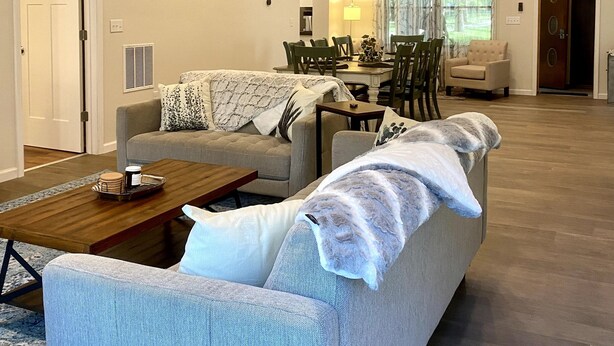 A spacious living room with a wooden ceiling, a grey sofa, a coffee table, and a dining area with chairs and a table. - Relax in our large 4b/2b ranch w/hot tub,firepit, game room, sleeps 11, pets ok (Sawyer)