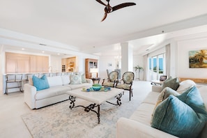A spacious living room with a white sofa, a coffee table, and two armchairs. A ceiling fan is visible, and there is a view of a balcony through large windows.
