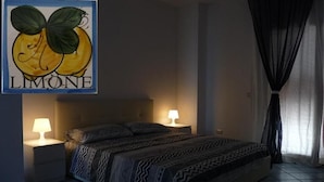Une chambre à coucher avec un lit, deux tables de chevet, une commode, une œuvre d’art représentant des citrons accrochée au mur, et une fenêtre avec des rideaux.