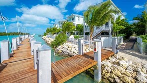 A wooden dock leading to a white building with a balcony, surrounded by palm trees and a clear blue sky.
