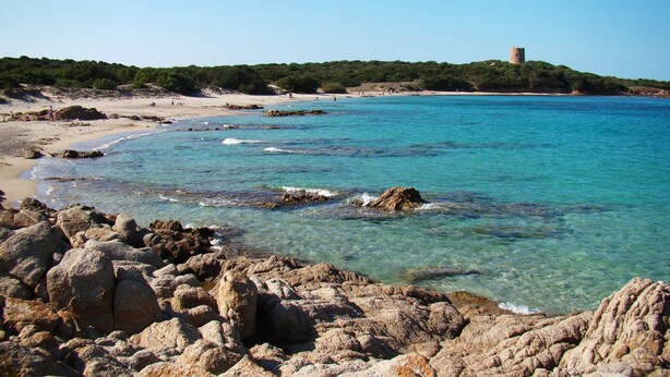 A coastal landscape with clear turquoise waters, rocky shorelines, and a distant castle on a hill.