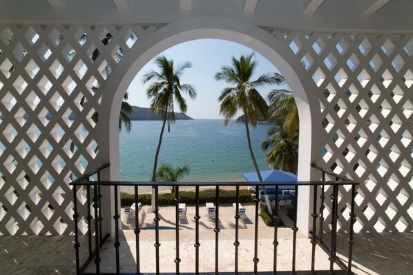 A view through a white lattice structure to a beach with palm trees and a pool area.