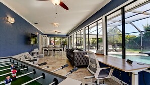 Ein geräumiges Zimmer mit Kicker, einer gemütlichen Couch, einem Schreibtisch mit Computer und großen Fenstern mit Blick auf einen Pool und eine Außenbestuhlung.