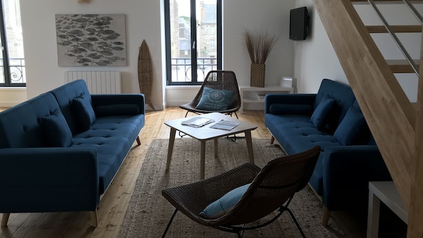 Een moderne woonkamer met twee blauwe banken, een rieten fauteuil, een salontafel en een groot raam met uitzicht op gebouwen.