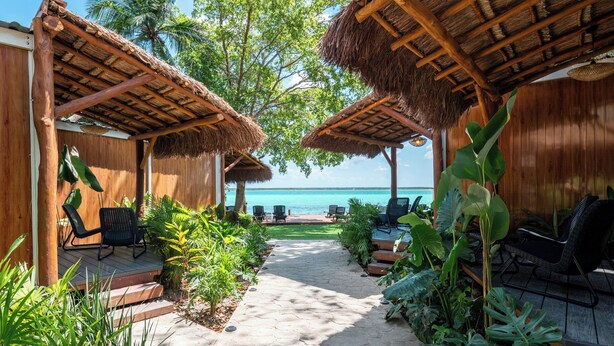 Una estructura de madera con techos de paja, asientos al aire libre y un sendero que conduce a una playa.
