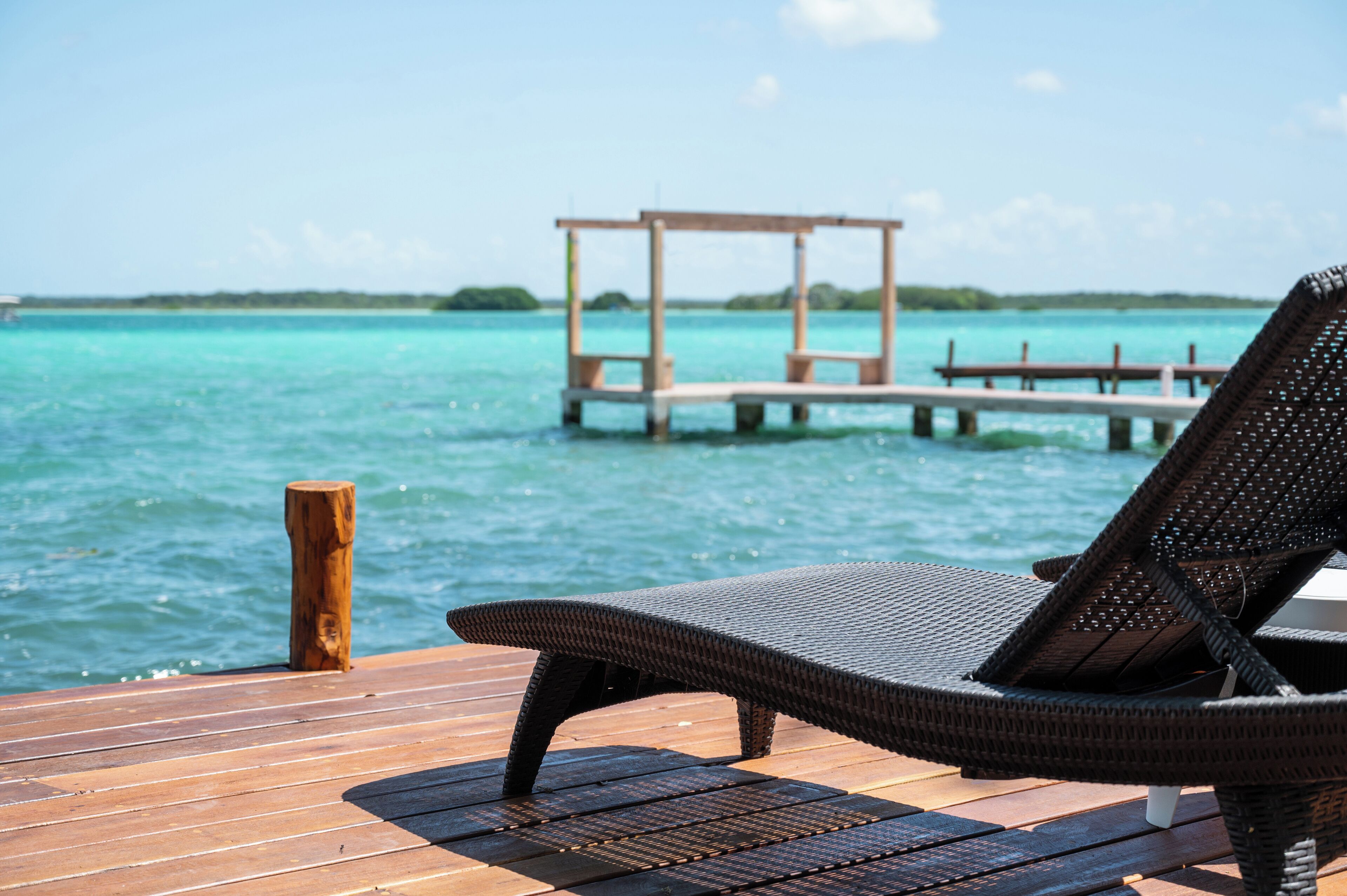 A wooden deck with a wicker lounge chair and a wooden pier extending into turquoise water.