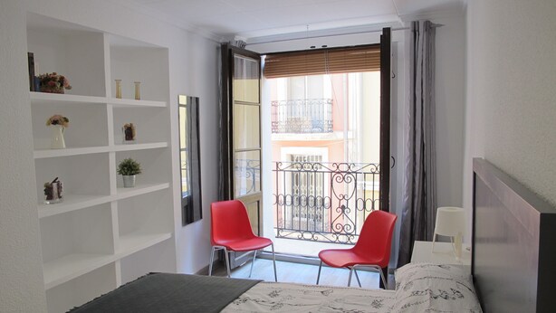 Une petite chambre à coucher, bien rangée, avec un lit, deux chaises rouges, une table de chevet blanche et une étagère ornée d’objets décoratifs.